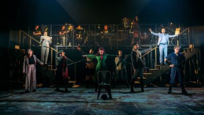 Actors dancing on stage in Urinetown, with the band visible on a raised level in the background